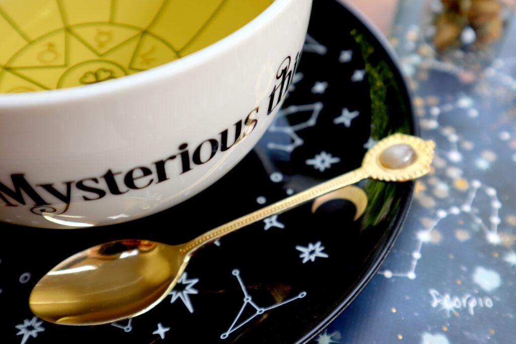 Closeup shot of a witchy white tasseomancy teacup containing herbal tea, a matching black saucer with stars and constellations, a vintage gold agate spoon, and a blue zodiac notebook.