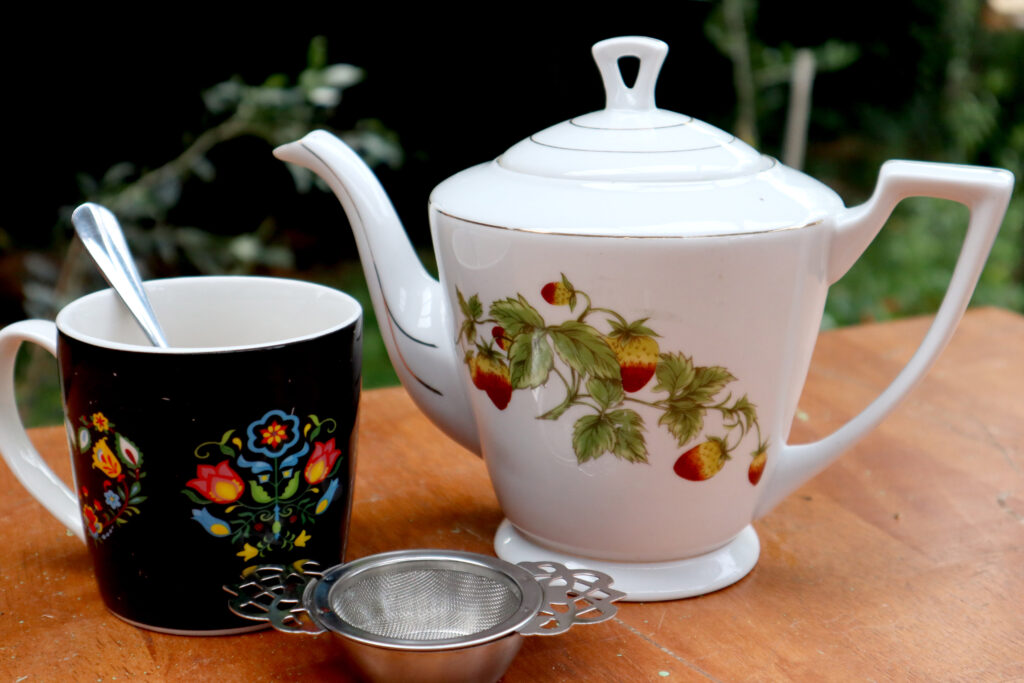 Image shpws a white Vintage ceramic teapot with a strawberries and leaves motif. The teapot sits on a timber surface, there is greenery in the background. There is also a black mug with a colourful folk art motif, with a spoon inside, and a vintage style tea strainer.