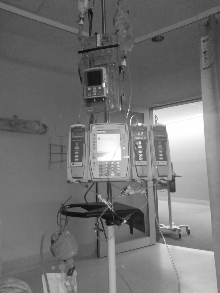 A black and white image of an IV pole in a dark hospital room. Hooked up to the IV pole are various bags and bottles of saline, medication, tubes, several different monitors, and drains for bodily fluids.