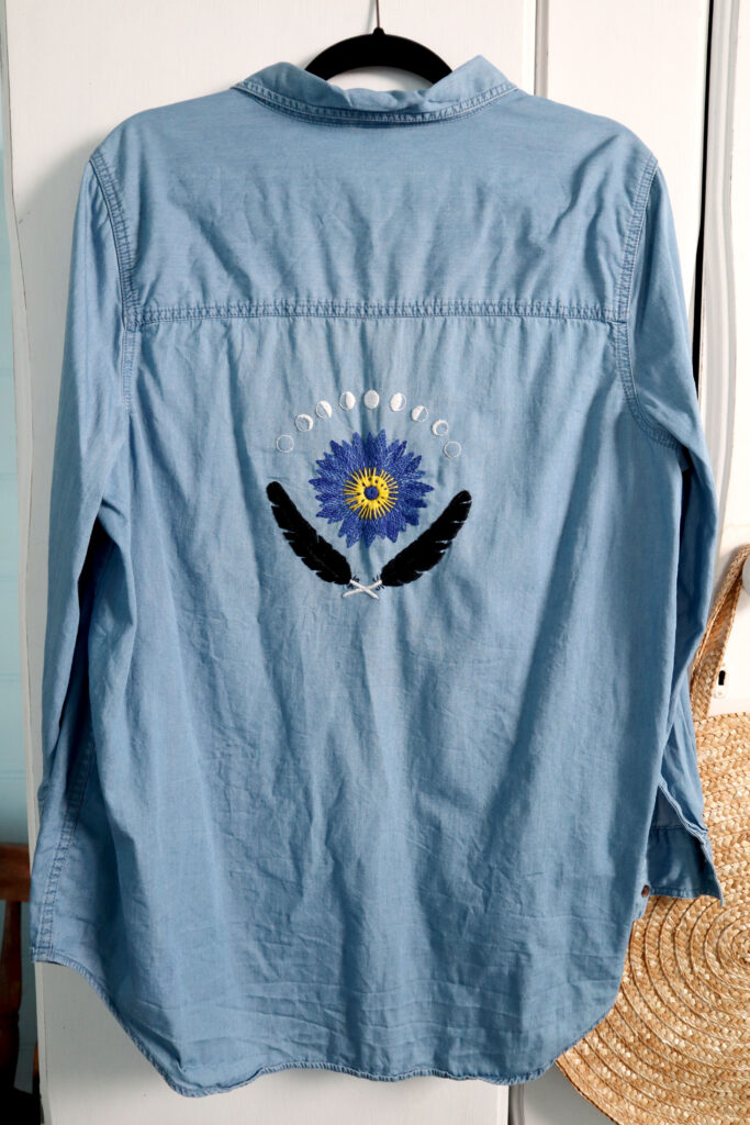 A blue chambray shirt hangs against the front of a vintage white wardrobe. the back of the shirt is shown. It is hand embroidered with black raven feathers, an egyptian blue lotus, and the phases of the moon. Behind the shirt hangs a round straw handbag.