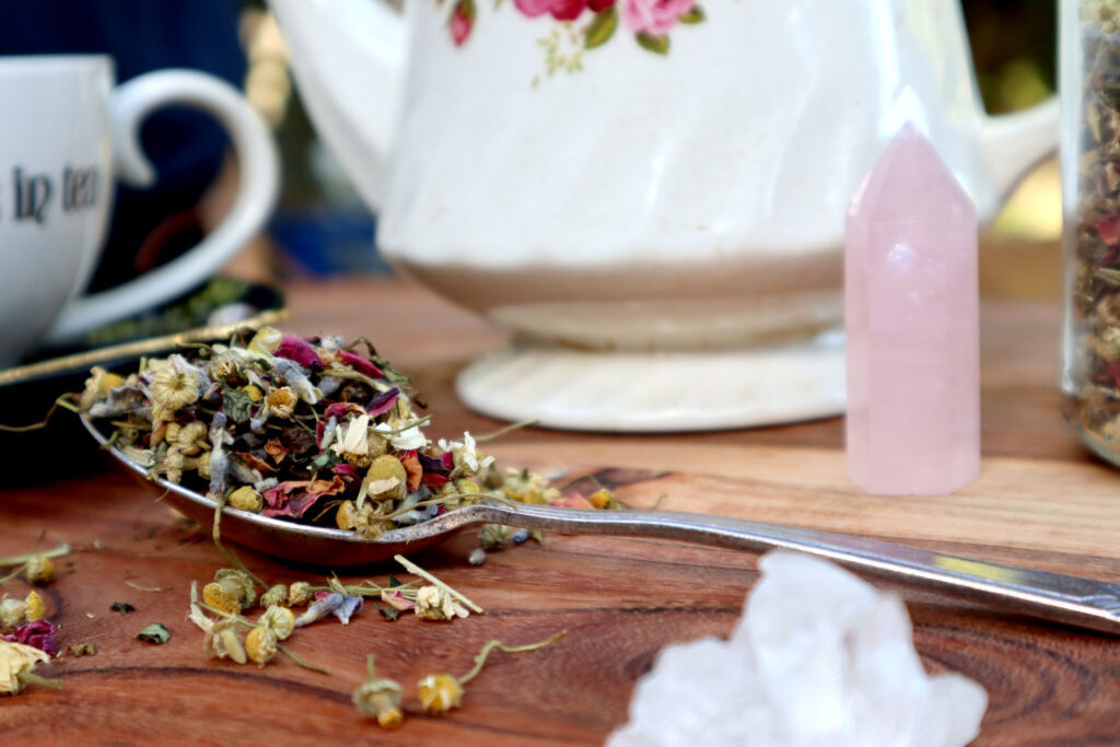 Uplifting Herbal tea Is displayed on a rustic timber surface. The shot is taken from a low angle close to the surface. Loose tea is spilled over a large vintage silver spoon. In the background are a witchy cup and saucer, a vintage gold agate teaspoon, a white vintage teapot, a glass jar of loose tea and a rose quartz tower. There is a raw himilayan quartz crystal in the foreground.