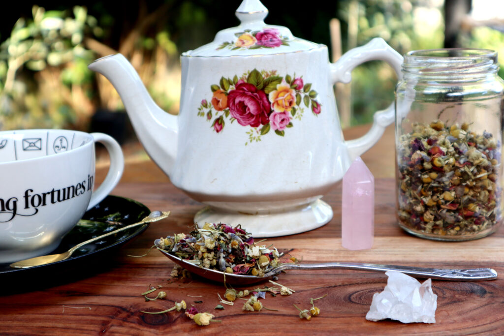 Uplifting Herbal Tea Is displayed on a rustic timber surface. Loose tea is spilled over a large vintage silver spoon. There is also a witchy cup and saucer full of brewed tea, a vintage gold teaspoon, a vintage rose pattern teapot, a glass jar of loose tea, a rose quartz tower, and a raw himilayan quartz crystal.