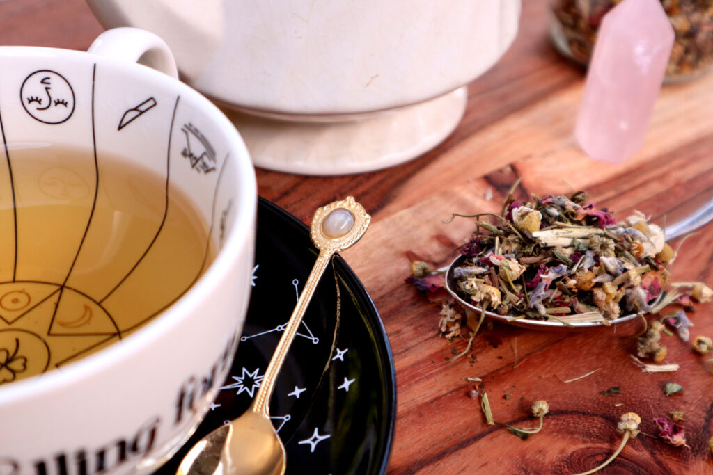 Uplifting Herbal tea Is displayed on a rustic timber surface. Loose tea is spilled over a large vintage silver spoon. There is also a witchy cup and saucer full of brewed tea, a vintage gold agate teaspoon, a vintage teapot, a glass jar of loose tea, and a rose quartz tower.