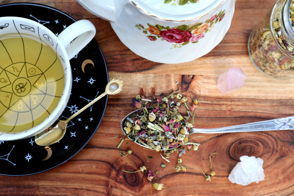 Uplifting Herbal Tea Is displayed on a rustic timber surface. The shot has been taken from above. Loose tea is spilled over a large vintage silver spoon. There is also a witchy cup and saucer full of brewed tea, a vintage gold teaspoon, a vintage rose pattern teapot, a glass jar of loose tea, a rose quartz tower, and a raw himilayan quartz crystal.