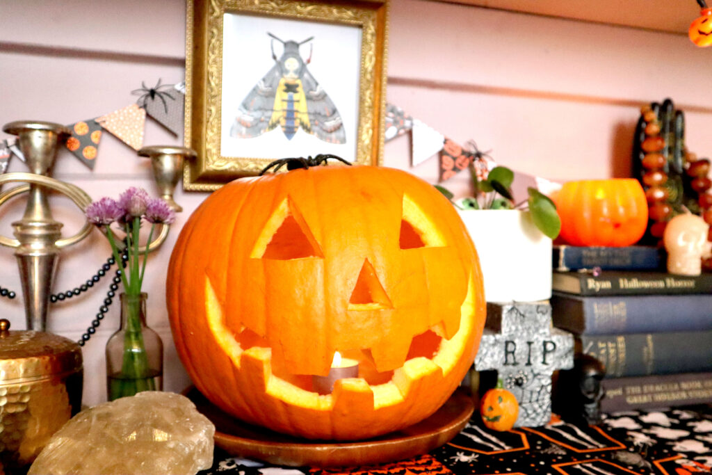 Image shows a Halloween display on top of a vintage halloween table runner. There is a carved jack o'lantern in the centre of the display. To the left is a silver candlestick draped with a strand of black pearls, a small vintage glass bottle containing purple chive flowers and a chunk of raw calcite crystal. To the left is a gravestone shaped candle, a small skull shaped candle, and an indoor plant. On top of a stack of vintage frabric-covered hardback horror stories, sits a jack o'lantern tealight holder, a small bone coloured skull candle, and a black resin palmistry hand draped with a vintage orange bakelite necklace.. On the wall behind these objects hangs mini triangle bunting and an original illustration of a death's head hawkmoth in a vintage gold square frame.